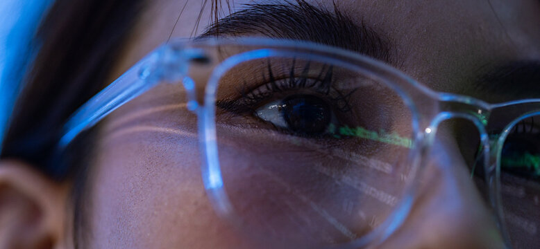 Close up eye, eyeglass, woman, reflection, blue light, dark hair, face detail show focused gaze through clear frame with eyelash and skin texture