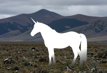 Fototapeta premium White unicorn silhouette standing in a dry, grassy field with mountains in the background under a cloudy sky, the background features a range of mountains under a cloudy sky