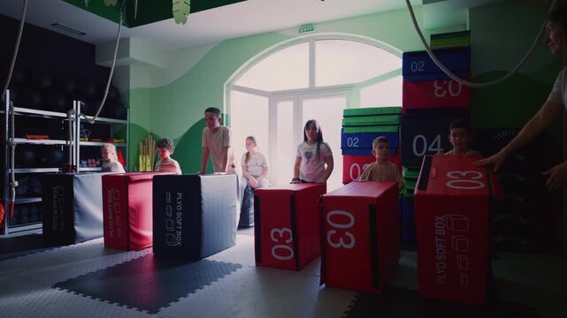 Kids participates in a dynamic exercise session, moving plyometric soft boxes under adult supervision. Children are actively training their bodies and developing strength in a modern gym environment.