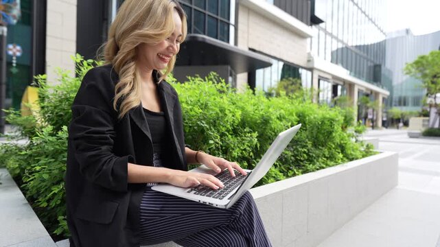 Asian businesswoman smiles while typing on her laptop seated on a garden ledge, reaching out with one hand before returning to type again in the Bangkok business district greenery.