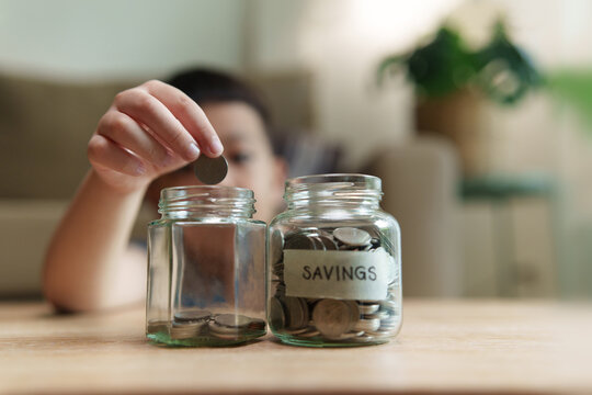 Asian Child placing a coin into a glass savings jar at home, representing money saving, budgeting, financial planning, education funds, savings goals and responsible financial habits.