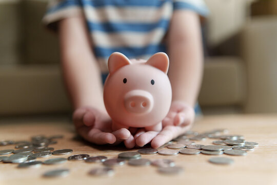Asian Child holding a pink piggy bank surrounded by coins at home, representing money saving, financial planning, budgeting, future investment, education funds and financial security.