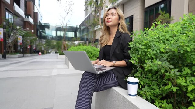 Asian businesswoman sits on a stone ledge typing on her laptop, lifting a coffee cup to sip before touching her shoulder, surrounded by greenery in the Bangkok business district.
