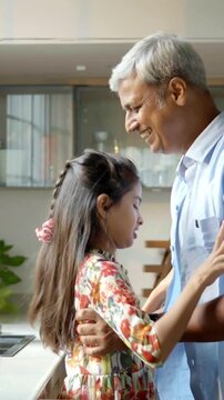 An Indian Asian father in his late forties playfully encourages his small daughter to drink milk while she sits happily on the kitchen platform, creating a joyful moment filled with love and laughter