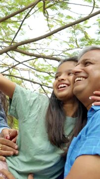 An Indian Asian family of four, with parents in their forties and two young kids, enjoying an outdoor moment while smiling and pointing at something in the distance, sharing happiness together