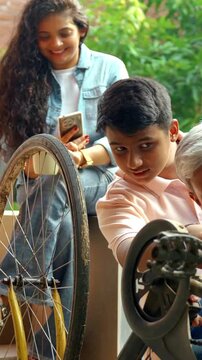 Indian Asian father repairing his child's bicycle on the house porch, with his son and daughter watching eagerly, while the wife looks on with a smile, capturing a happy family moment