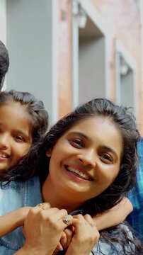 An Indian Asian family of four, parents in their forties and two kids, smiling while posing for a joyful portrait outside their lavish home, capturing a cherished moment together.