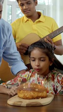 An Indian Asian family of four joyfully celebrates their small daughter's birthday by cutting a cake adorned with candles, surrounded by glitter, love, and laughter as they pose for a memorable photo