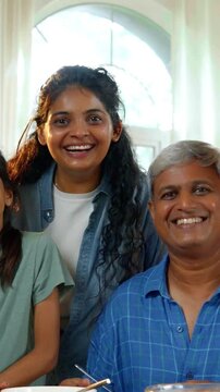An Indian Asian family of four, with parents in their forties, their teenage son and small daughter, joyfully posing for a photo at the dining table filled with delicious food, showing happy moments