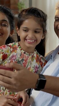 Indian Asian family of four, with parents in their forties and two kids, sitting closely on a sofa laughing out loud while enjoying a funny video on a smartphone, sharing a joyful moment together
