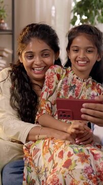 Indian Asian family of four, with parents in their forties and two kids, sitting closely on a sofa laughing out loud while enjoying a funny video on a smartphone, sharing a joyful moment together