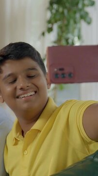 A closeup of a joyful Indian Asian family, with parents in their mid-forties, a teenage son, and a small daughter sitting together on the sofa while the wife captures a selfie with her smartphone