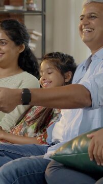 Indian Asian family of four, parents in their forties and two kids, sitting closely on a sofa, laughing out loud while watching television together, enjoying a fun-filled family evening at home