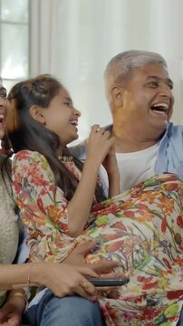 Indian Asian family of four, parents in their forties and two kids, sitting closely on a sofa, laughing out loud while watching television together, enjoying a fun-filled family evening at home