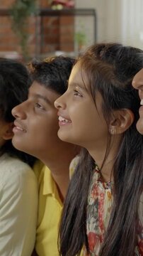 Indian Asian family of four, parents in their forties, sitting closely on a sofa with their two kids, smiling warmly while looking at the camera, capturing a joyful moment together indoors