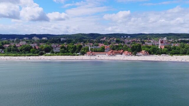 Aerial view of the sandy beach and coastline of Sopot in Poland, showing people relaxing, buildings with red roofs, and calm sea water.