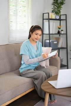 Young Asian female doctor writing notes on clipboard while sitting on sofa in modern home office
