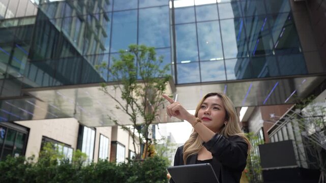 Businesswoman stands beneath a glass skywalk holding a tablet and stylus, tilting her chin upward to study the architecture as the static camera frames the towers overhead.