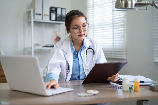 Young Asian female doctor reviewing medical records on tablet and laptop in modern clinic office