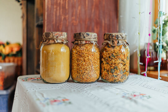 Three glass jars filled with golden dried flower petals, herbal material and fine yellow powder displayed on an embroidered tablecloth in a cozy home interior