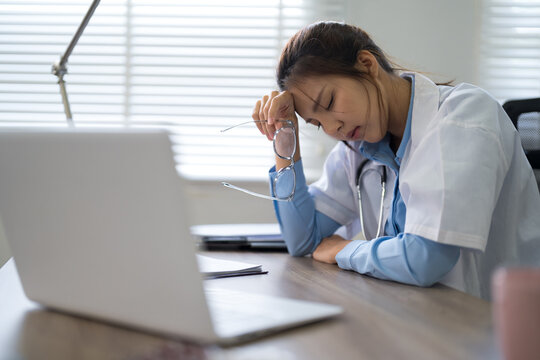 Exhausted Asian female doctor experiencing burnout and stress while working at laptop in modern hospital office