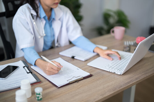 Asian female doctor writing patient notes on clipboard while using laptop in modern clinic office with copy space