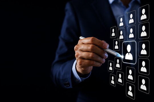 A businessman in a dark suit using a stylus to select a human from a digital grid of user profile symbols on a black background screen