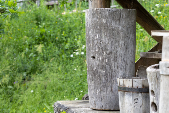 A traditional hollowed tree trunk serves as a wooden beehive. The object stands outdoors on a wooden step near timber buckets, set against a blurred background of green summer grass.