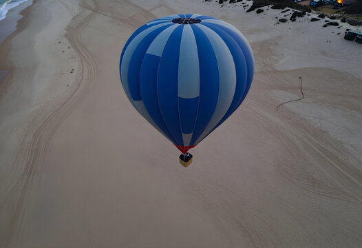 Blue and white striped hot air balloon flying over a sandy beach with Tire tracks and ocean waves, Tire tracks crisscross the sand, leading towards the distant ocean waves
