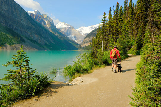 Man in Red Shirt Walking Dog on Leash on Mountain Trail Next to Blue Lake in Wilderness for Outdoor Adventure