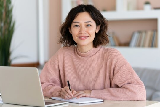 Young Asian woman sitting at a desk with a laptop, holding a pen and notebook, smiling while participating in an online meeting in a modern home office setting