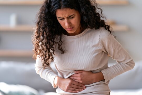 Young woman with voluminous curly dark hair holds her stomach in discomfort while sitting on a couch in a modern living room with neutral tones and soft furnishings