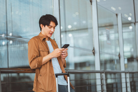 Young man using smartphone at airport with luggage, waiting for flight and checking travel information