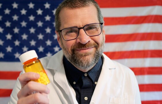 Pharmacist in lab coat holding prescription pill bottle, cash, and crypto against an American flag, symbolizing the influence of money on US medicine