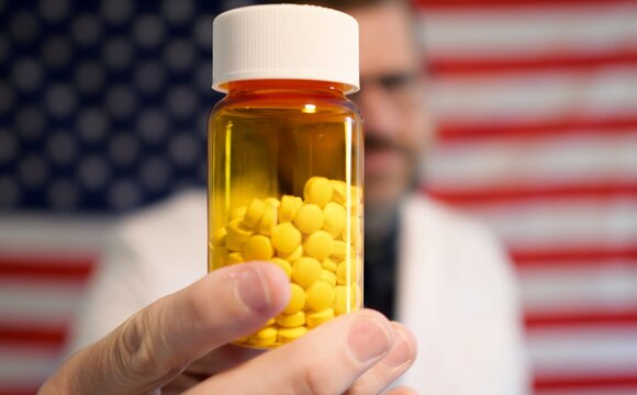 Pharmacist in lab coat holding prescription pill bottle, cash, and crypto against an American flag, symbolizing the influence of money on US medicine