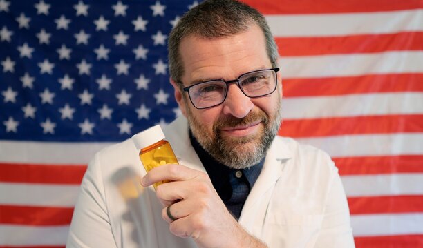 Pharmacist in lab coat holding prescription pill bottle, cash, and crypto against an American flag, symbolizing the influence of money on US medicine