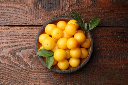 Fresh yellow cherry plums with green leaves in bowl on wooden table, top view