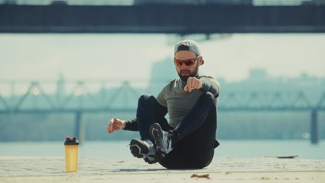 Athlete Sitting On Ground Stretching Leg Muscles Before Workout. Sports Training Near River Bridge
