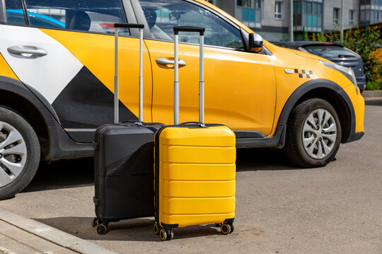 Two suitcases, one black and one yellow, positioned beside a parked yellow and black taxi car on a city street with modern buildings in the background