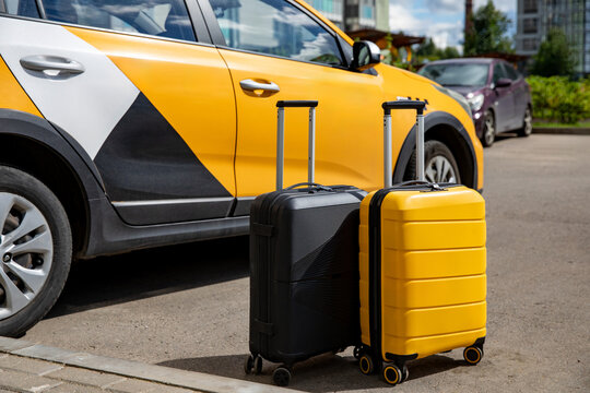 Two suitcases, one black and one yellow, positioned beside a parked yellow and black taxi car on a city street with modern buildings in the background