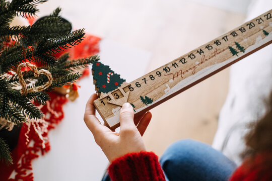 Woman hand holds wooden MERRY CHRISTMAS countdown calendar with sliding tree marker near decorated tree. Wooden christmas countdown, advent countdown calendar, handmade advent calendar