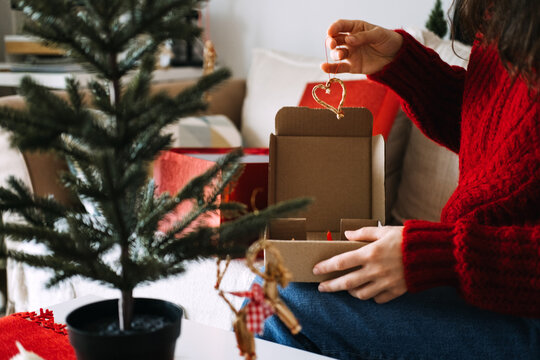 Woman hands untie ribbon on straw ornament above box of decorations on lap. Sustainable christmas decorations, natural material ornaments, eco conscious holiday decor