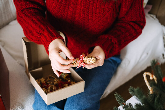 Woman hands untie ribbon on straw ornament above box of decorations on lap. Sustainable christmas decorations, natural material ornaments, eco conscious holiday decor