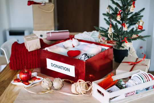 DONATION box filled with clothes stands on table beside Christmas tree and wrapped gifts. Christmas donation box, holiday charity clothing drive, donating warm clothes winter