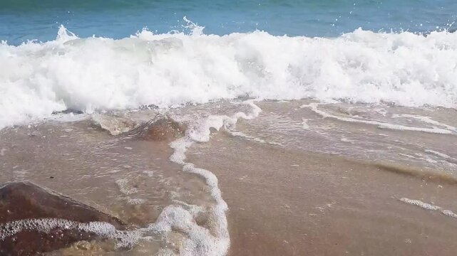 Close-up of foamy coastal surf with azure stormy sea waves rolling on sand beach and wet rocks on foreground at sunny summer day on Baltic sea shore. No people seaside water surface natural background