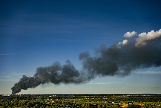 Black smoke billowing from a warehouse fire creating a dark plume against the blue sky, impacting air quality over the urban landscape and surrounding green environment