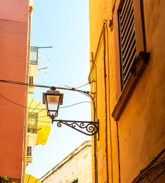 Narrow alley in Bari, Italy with warm yellow and orange building facades, ornate wrought iron street lamp mounted on wall, wooden shutters and blue sky above