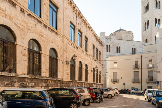 Bari Italy old town street with historic limestone palace facade featuring arched windows, parked cars on cobblestone square near Bari Cathedral bell tower