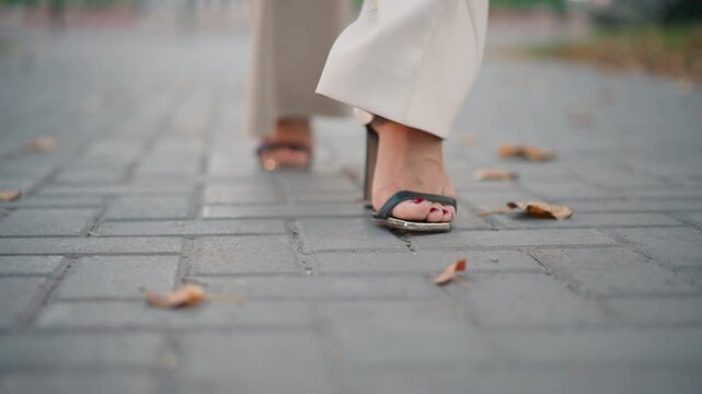 Ground level view shows feet wearing open toe heeled sandals moving across concrete pavers, with one foot landing and other lifting in steady forward motion under soft daylight, with sandal, heel
