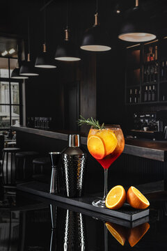 Close-up of Aperol on bar counter. Tall glass holds bright orange drink with ice and orange slices. Stylish black shaker with textured surface and measuring cup sit nearby. There's plenty of dark, emp